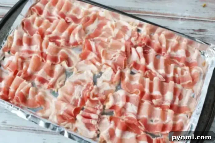 A baking tray lined with aluminum foil is covered with neatly arranged strips of raw bacon, ready to be cooked in the oven for a delicious, crispy result.