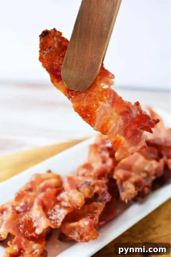A piece of crispy bacon held by wooden tongs above a white rectangular plate filled with more bacon strips. The background is blurred, and the focus is on the bacon being lifted, showcasing perfectly cooked strips—just like what you'd get if you knew how to cook bacon in the oven with aluminum foil.