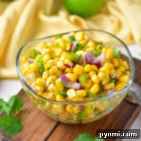 A glass bowl of roasted chili corn salsa garnished with red onion and herbs rests on a wooden board.