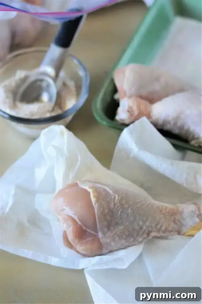 Oven-Baked Chicken Drumsticks 3 Raw chicken drumsticks are on white paper towels, ready for preparation. A small scoop rests in a bowl nearby containing a flour mixture. A green tray in the background holds additional chicken drumsticks, soon to become baked chicken legs.