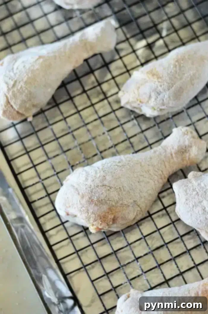 Oven-Baked Chicken Drumsticks 5 Baked chicken legs are coated in flour and placed on a wire rack for drying. The rack sits on a baking sheet lined with foil, suggesting the drumsticks are being prepared for frying or baking.
