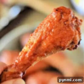 A hand holding a crispy, saucy baked chicken leg, reminiscent of buffalo chicken drumsticks, with a blurred background.