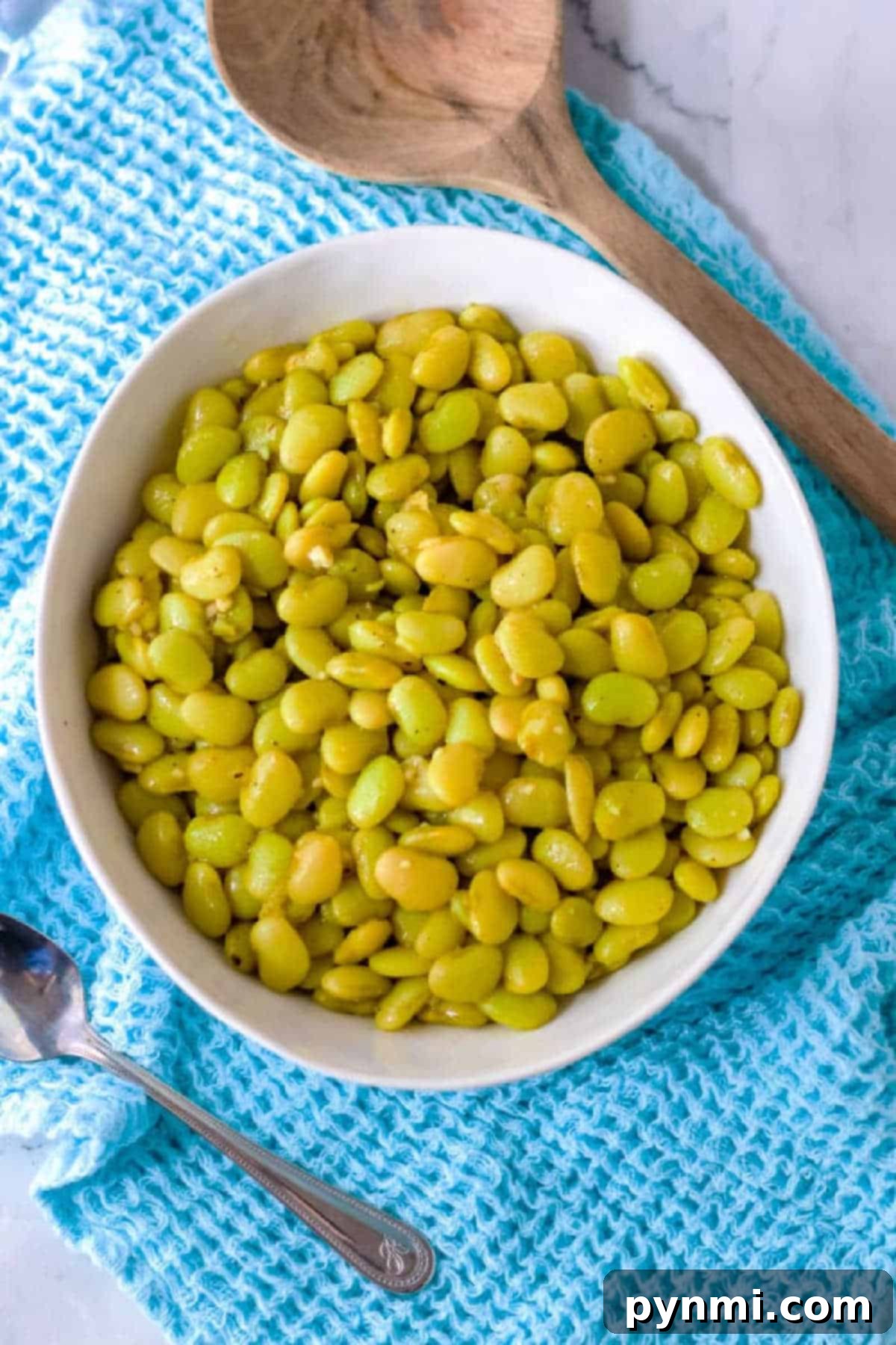 A bowl of perfectly cooked, seasoned dried lima beans, ready to be served, sitting on a blue cloth next to a wooden spoon.