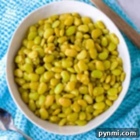A bowl of cooked dried lima beans sits on a blue cloth, with a wooden spoon and metal spoon nearby.