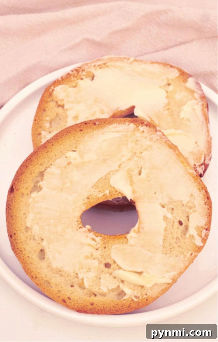 Two buttered bagel halves rest on a white plate atop a light-colored fabric background, tempting you to learn how to toast a bagel in the oven to perfection.