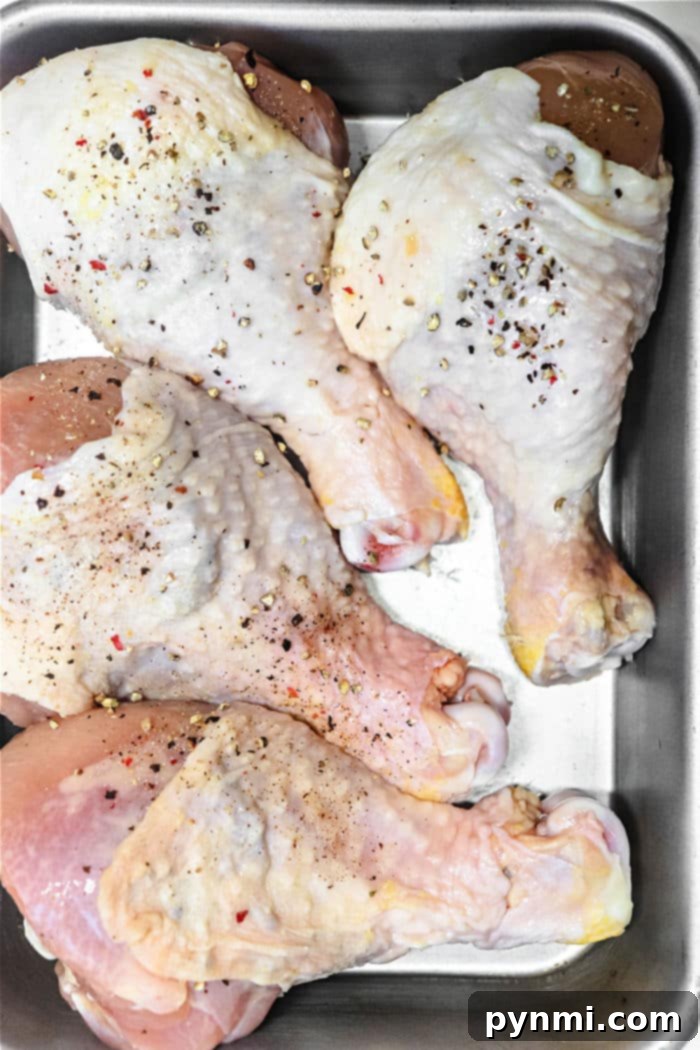 Four raw chicken legs in a metal baking dish, seasoned with salt, pepper, and spices, ready to transform into smoky chicken drumsticks.