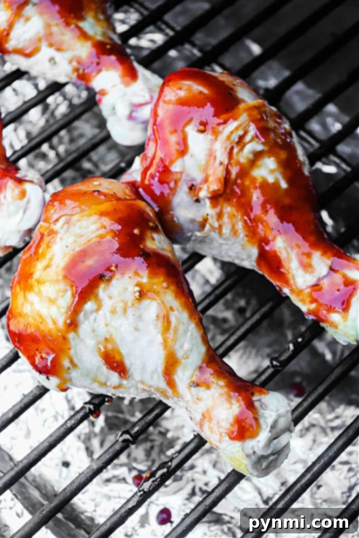 Smoked chicken drumsticks brushed with red sauce sizzle on the grill. Foil beneath catches drips as the glossy glaze coats each piece, enhancing their mouthwatering appeal.