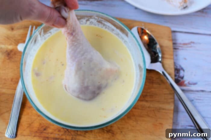 A hand dipping a raw chicken leg into a creamy buttermilk and egg batter, preparing it for frying.