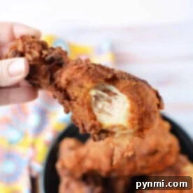 A close-up captures a hand holding a fried chicken drumstick with a bite taken out, hovering over a bowl full of crispy fried chicken legs.