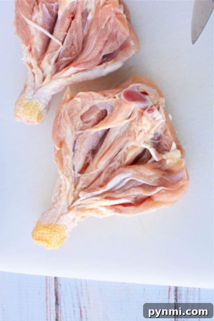 Two raw, butterflied chicken legs rest on a white cutting board, partially cut open, with a knife visible in the top right corner, illustrating the butterflying technique with precision.
