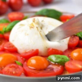 A knife slices through the creamy burrata in this Burrata Caprese dish, surrounded by juicy cherry tomatoes and fresh basil on a plate.