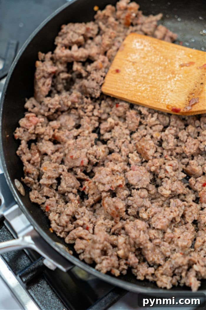 Creamy Velveeta Sausage Rotel Dip 8 Ground sausage sizzling in a black skillet on the stovetop, stirred gently with a wooden spatula, forming a savory base for a hearty Velveeta and Rotel dip.