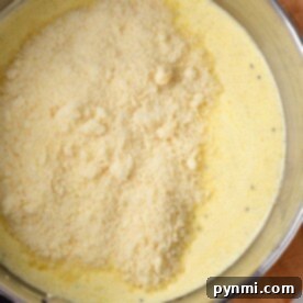 A pot of creamy yellow Cajun Alfredo sauce, topped with a pile of grated cheese, rests invitingly on a wooden board.