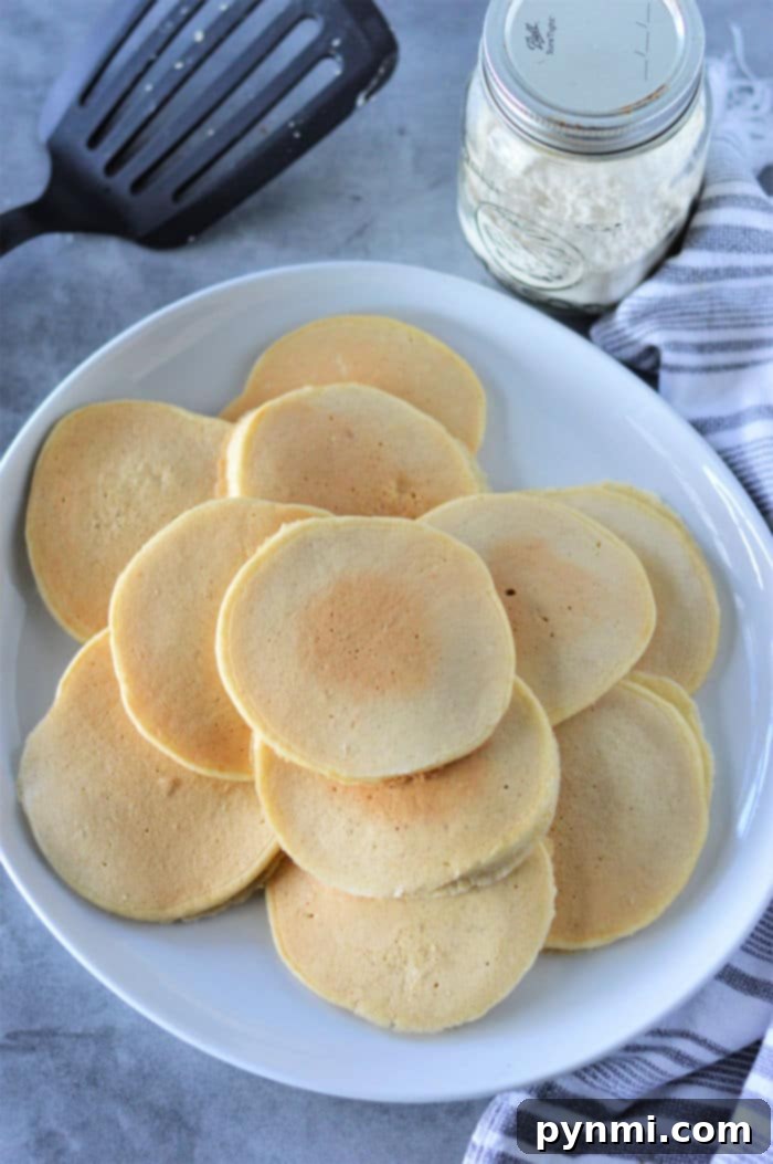 Cracker Barrel-Inspired Homemade Pancake Mix 5 A perfectly arranged stack of round, fluffy Cracker Barrel Pancake Mix pancakes on a white plate, accompanied by a spatula and a jar of flour in the background, all resting on a striped napkin.