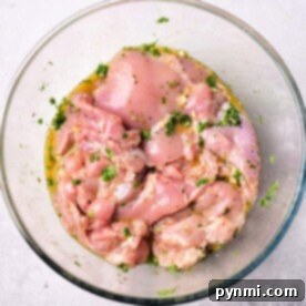 Raw, marinated chicken pieces, infused with a Mediterranean chicken marinade and herbs, rest in a clear glass bowl on a light surface.