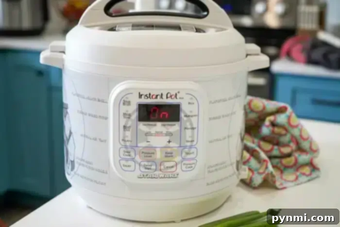 A white Instant Pot with a Star Wars theme sits on the kitchen counter, ready to tackle your favorite beef arm roast recipe. The digital display glows "On." Next to it lie a patterned cloth and some green onions, all against the backdrop of blue kitchen cabinets and a stovetop.