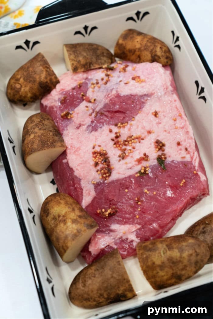 A raw corned beef brisket, seasoned, nestled in a rectangular baking dish with halved potatoes arranged around it. The dish, with its classic white interior, illustrates a straightforward way to cook corned beef in the oven.