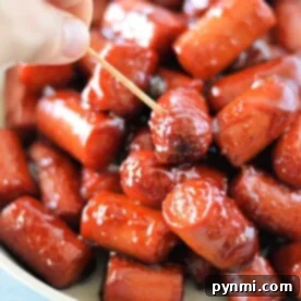 A hand holding a toothpick picks up a glazed mini sausage from a bowl of hot dog burnt ends.