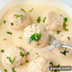 A bowl of creamy white sauce with meatballs, garnished with chopped herbs. A spoon lifts one Alfredo meatball, enticing you to savor its rich flavor.