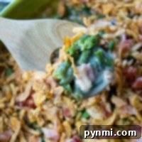 A wooden spoon lifts a serving of easy green bean casserole with crispy onions and diced ham from a green baking dish.