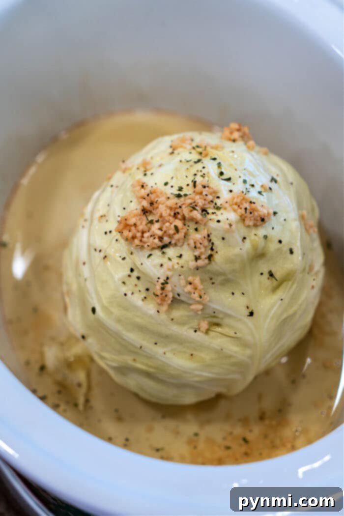 Perfect Slow Cooker Whole Cabbage 5 A whole head of cabbage, slowly cooked to perfection in a white dish, is beautifully topped with minced garlic and herbs nestled in a savory broth.