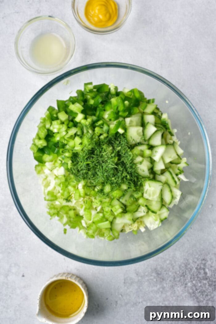 Effortless Asian Cabbage Slaw 3 Bowl of chopped green vegetables and herbs, including cabbage, cucumber, and bell peppers, ready for dressing. Small bowls of mustard, lemon juice, and olive oil are arranged neatly beside it.