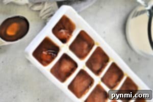 A white ice cube tray filled with brown liquid, presumably frozen coffee, is placed on a light gray countertop. To the left of the tray, a teaspoon holds a small amount of the same brown liquid. A gray cloth and a glass pitcher are partially visible in the background, hinting at how to make cold foam for coffee.