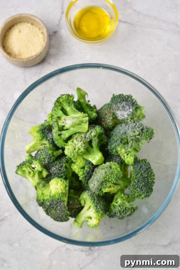 A glass bowl with frozen broccoli florets, ready for seasoning, sits next to small bowls of grated cheese and olive oil on a marble surface, highlighting the minimal ingredients needed for baking.