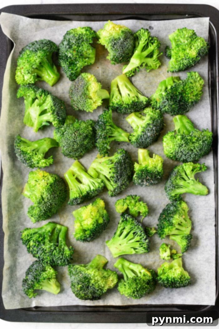 Frozen broccoli florets coated in olive oil and seasonings, spread in a single layer on parchment paper on a baking sheet, ready for roasting.