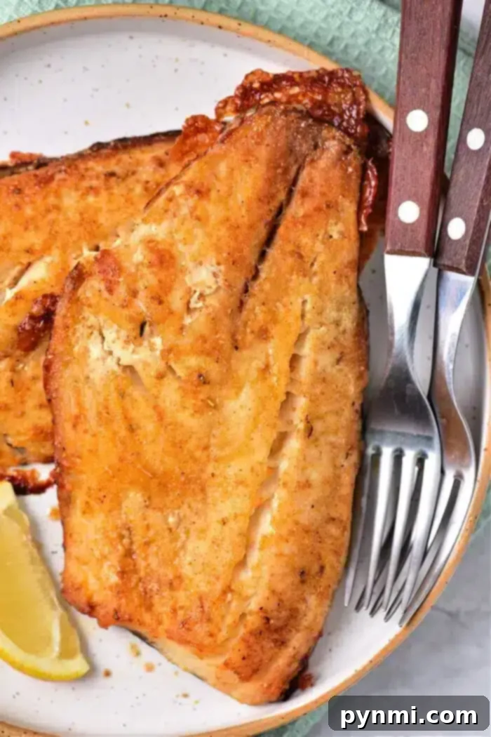 Grilled fish fillets with a golden crust rest on a white plate, alongside a lemon wedge. Perfectly cooked, just like baking frozen fish to perfection. A set of silverware with wooden handles is placed beside them, with a green textured cloth partially visible in the background.