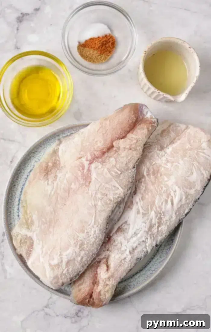 Two raw fish fillets sit on a plate, surrounded by small bowls containing olive oil, seasoning mix, and a light-colored liquid on a marble surface—ideal ingredients when baking frozen fish to perfection.