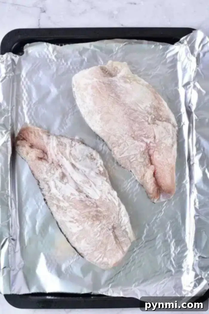 Two raw fish fillets rest on a baking sheet lined with aluminum foil, ready for baking. Their light, frosty appearance suggests they're frozen. The simple, light background enhances their fresh allure.