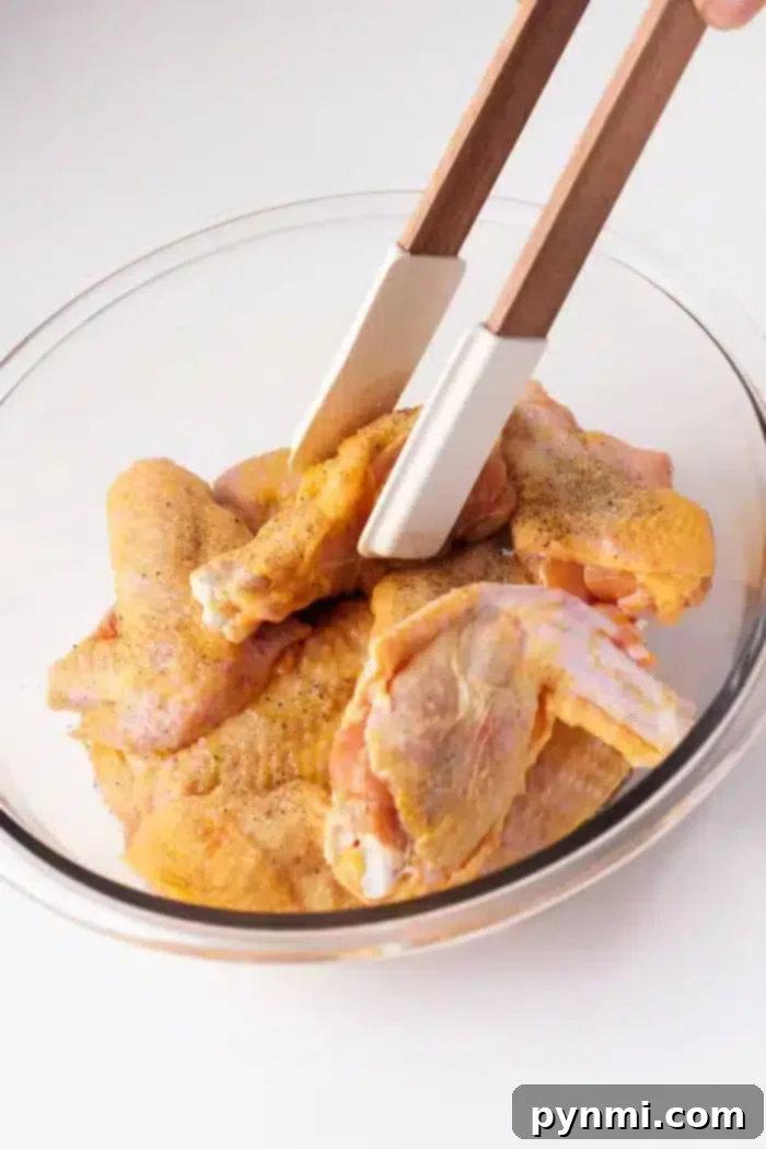 A glass bowl contains several raw chicken wings seasoned with spices.