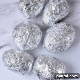 Six potatoes wrapped in aluminum foil rest on a marble surface, ready to answer the question: how long to bake potatoes at 375 in foil?.