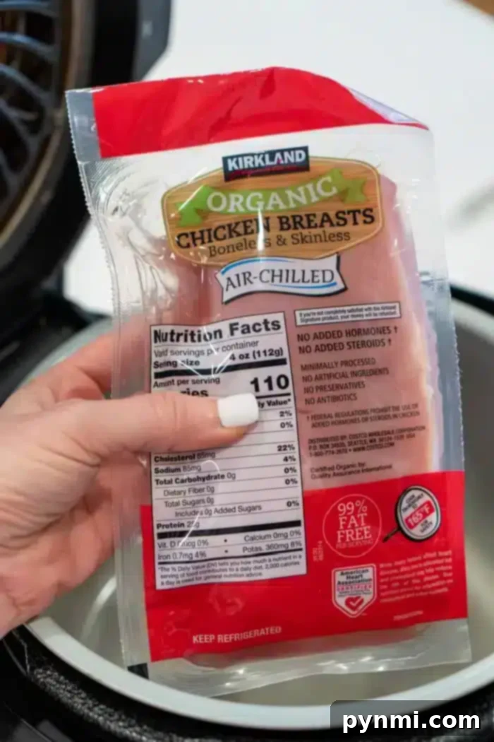 Perfect Ninja Foodi Shredded Chicken 4 A hand holds a package of Kirkland organic chicken breasts over a Ninja Foodi, preparing to cook delicious shredded chicken. The boneless, skinless, air-chilled packaging displays nutritional facts and other important details for conscious cooking.