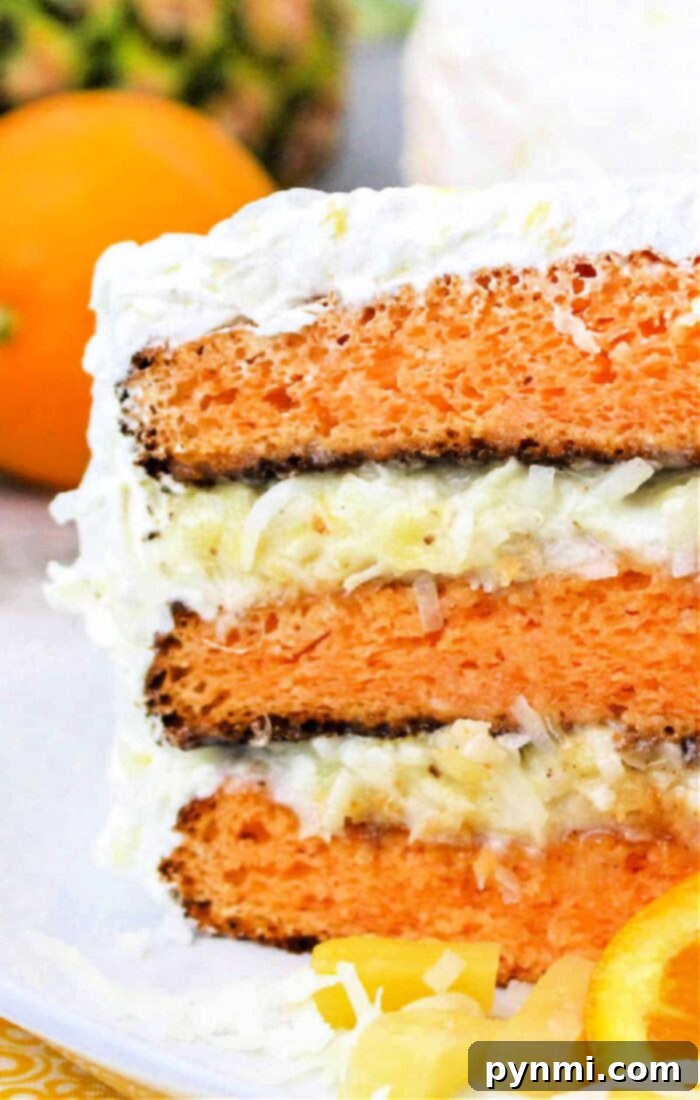 Aloha Bliss Cake 8 A close-up of a slice of layered orange and white Hawaiian cake with frosting, featuring visible layers of orange cake, white frosting, and shredded coconut. There is an orange and some pineapple pieces in the background, hinting at its tropical flavors.