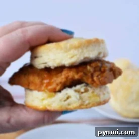 A hand grasps a delicious Whataburger Honey Butter Chicken Biscuit, with its golden layers tempting the eye, while a plate and another biscuit wait invitingly in the background.