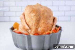 A whole seasoned chicken sits upright in a bundt pan, surrounded by perfectly sliced potatoes and carrots. Wondering how long to cook a whole chicken like this? The backdrop is a simple white tile wall, adding an elegant touch.