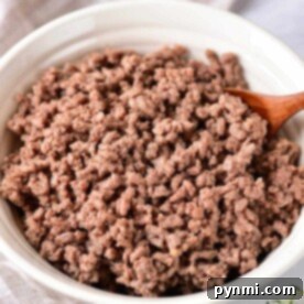 Cooked ground beef in a white bowl with a wooden spoon rests on a cloth napkin, embodying the perfect texture achieved by knowing how long to cook ground meat just right.