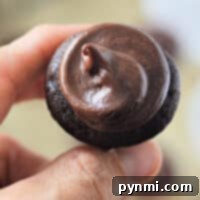 A hand holding a mini chocolate cupcake with thick chocolate frosting on top.