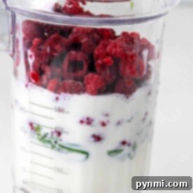 A blender jar filled with milk, frozen raspberries, and greens is prepped and ready to show you how to make a thick smoothie.