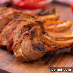 Grilled, seasoned lamb ribs rest on a wooden board, accompanied by a bowl of red dipping sauce in the background—an inviting glimpse into how to cook lamb ribs to perfection.