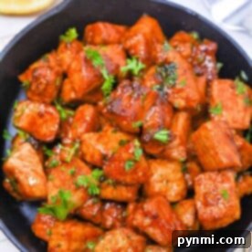 Pork bites, glazed with a savory sauce in a skillet, are garnished with fresh herbs and served alongside lemon slices, with forks at the ready for easy dining.