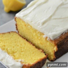 Effortless Lemon Pound Cake with Cake Mix 6 A frosted lemon pound cake with a slice cut, perched invitingly on a white plate, hints at its delightful origin using cake mix. A whole lemon sits artfully in the background, adding a touch of zest to the scene.