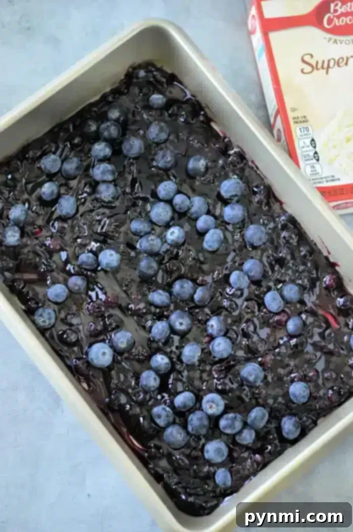 A baking pan brimming with blueberry pie filling, topped with fresh blueberries, showcases a delectable blueberry cobbler made with Betty Crocker Super Moist white cake mix. The partially visible box hints at the delightful blend that elevates this dessert on the gray surface.