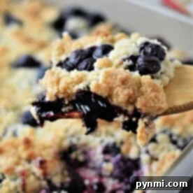 A portion of blueberry cobbler, expertly crafted with cake mix, is being served from a baking dish using a wooden spoon.