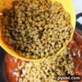 A hand demonstrating how to cook green lentils on the stove, pouring them from a yellow strainer into a pot of sauce with a wooden spoon.