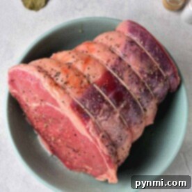A bowl showcasing a seasoned raw beef roast with visible spices, ready for its transformation into a succulent dish. Accompanied by aromatic bay leaves and a pepper shaker, it sets the stage for those eager to discover how to cook a roast perfectly.