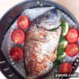 Air fryer whole fish seasoned with herbs and layered with sliced tomatoes rests in the basket, ready for cooking perfection.