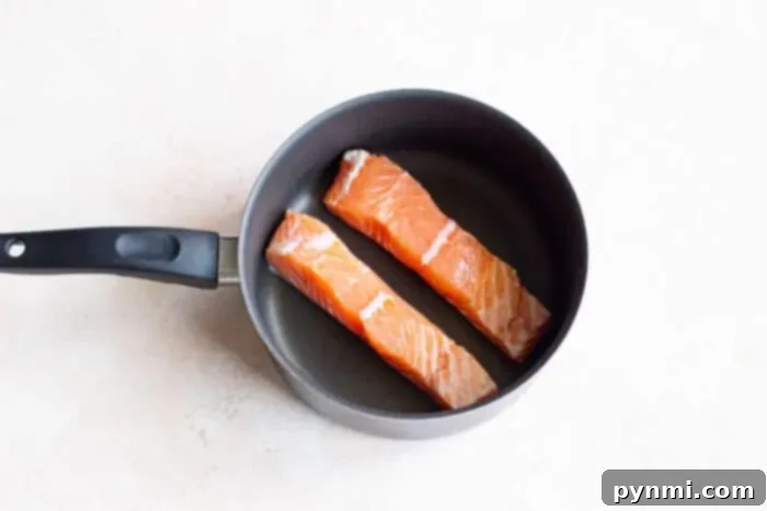 Delicate Poached Fish 3 Two raw salmon fillets prepared in a non-stick pan with a black handle, ready for gentle cooking in a flavorful broth on a light-colored surface.